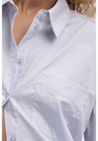 Light blue button-up shirt with a pointed collar, front pocket, and smooth texture. Features clear buttons and a fitted design.
