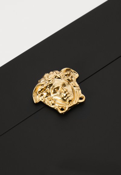 Melna virsma ar zelta dekoratīvu aizdares elementu, kas attēlo detalizētu sejas reljefu un sarežģītu matu dizainu, redzamas trīs pievienošanas caurumi.