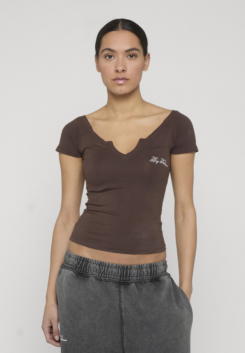 Νέα γυναίκα με σκούρα μαλλιά πιασμένα πίσω, φοράει στενό καφέ τοπ με V λαιμόκοψη και κοντά μανίκια, καθώς και άνετα γκρι παντελόνια φόρμας, στέκεται μπροστά σε ουδέτερο φόντο.