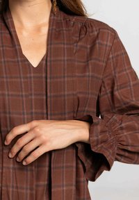 Blusa de cuadros marrones con escote en V, mangas abullonadas y fruncidos en los hombros. La tela parece suave con un acabado texturizado.