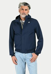 Giacca blu navy con chiusura a cerniera,'orlo e polsini a costine, e un logo sul petto, indossata sopra una maglietta grigia chiara e jeans.