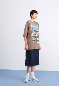 Brun oversize t-shirt med grafisk design, kombineret med en mørk denim blyantskirt. Modellen bærer hvide sokker og sølvfarvede sneakers.