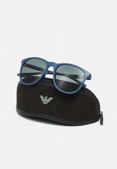 Occhiali da sole blu con finitura opaca all'interno di una custodia soft nera con un logo sottile e chiusura a zip, che mostrano una forma arrotondata e lenti scure.