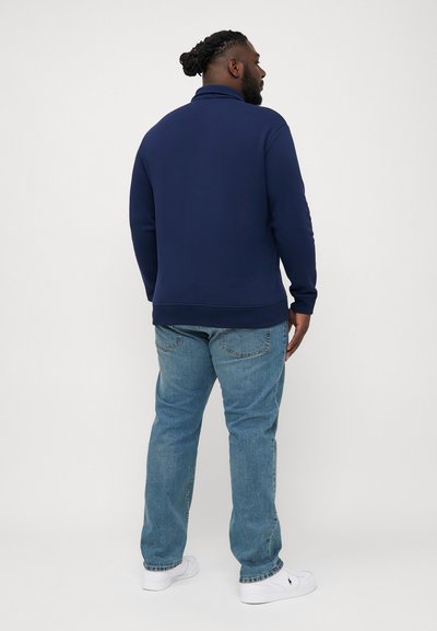 Marineblauwe pullover met hoge kraag, gecombineerd met lichtblauwe jeans. De pullover heeft lange mouwen en een relaxte pasvorm.