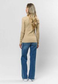 Beige, hosszú ujjú felső ribbed textúrával, kék farmer, bőszárú nadrággal párosítva. A modell enyhén az oldalára fordul, látható, hogy leengedett hajjal áll.