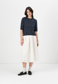 Navy cable-knit short-sleeve sweater with bead accents, paired with a white pleated midi skirt and black loafers over white socks.