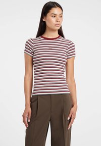 Donna che indossa un top aderente a maniche corte a righe rosse, bianche e nere e pantaloni a vita alta marroni con gamba larga, su uno sfondo bianco.