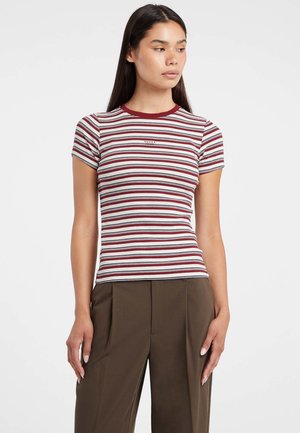 Donna che indossa un top aderente a maniche corte a righe rosse, bianche e nere e pantaloni a vita alta marroni con gamba larga, su uno sfondo bianco.