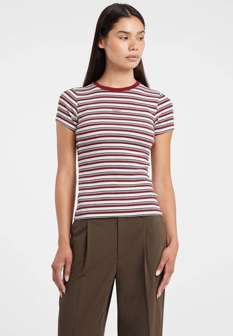 Donna che indossa un top aderente a maniche corte a righe rosse, bianche e nere e pantaloni a vita alta marroni con gamba larga, su uno sfondo bianco.