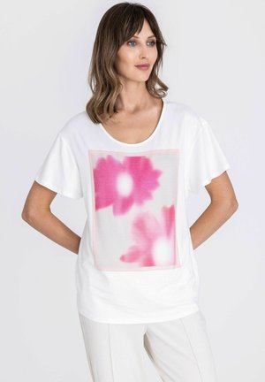 Vrouw draagt wit T-shirt met grote roze bloemmotief aan de voorkant, gecombineerd met witte broek, poseert met handen achter de rug.
