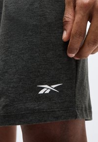 Κοντινό πλάνο ενός σκούρου γκρι ρούχου με λευκό λογότυπο Reebok κοντά στο τελείωμα και το χέρι ενός ατόμου που ακουμπά δίπλα του.