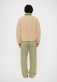 Beige fleece jack met hoge kraag, achteraanzicht. Gecombineerd met lichtgroene wijdvallende broek met zakken en gestikte details.