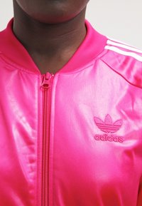 Rosa satin bomberjacka med dragkedja och broderad adidas-logga på vänster bröst. Randiga detaljer på ärmarna.