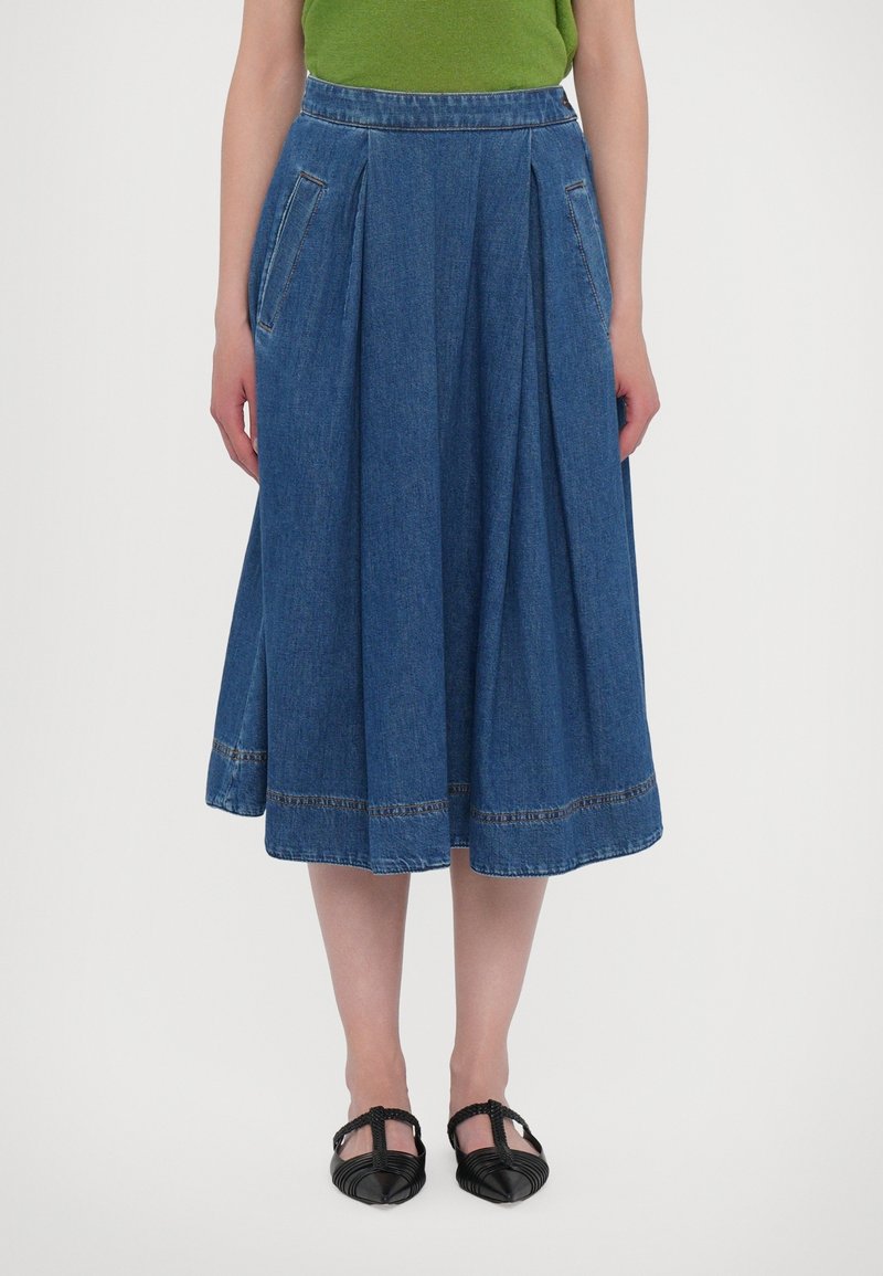 Gonna di jeans in una profonda tonalità di blu con pieghe, tasche laterali e un orlo caratterizzato da cuciture a contrasto. Indossata con ballerine nere texture.