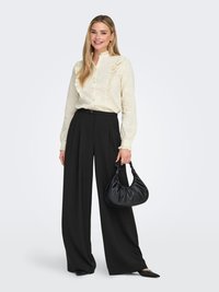 Crèmekleurige blouse met ruches en knoopsluiting, gecombineerd met zwarte, hooggetailleerde wijde broek. Model draagt een zwarte, zachte handtas.