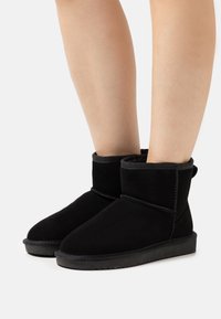 Bottines en suède noir avec un bout arrondi, doublure douce et coutures le long des seams. Semelle en caoutchouc basse avec une adhérence texturée.