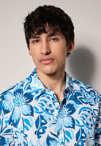 Chemise à manches courtes, boutonnée, blanche avec un motif floral bleu. Tissu doux et col standard.