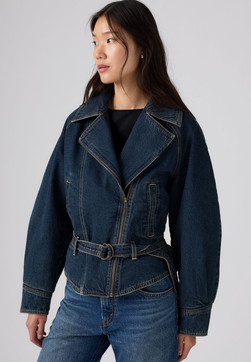 Femme aux longs cheveux foncés portant une veste en jean sombre ceinturée et un jean bleu, regardant vers sa gauche sur un fond uni.