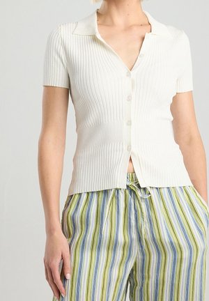 Kvinde iført en hvid ribbet kortærmet skjorte med knapper og løse bukser med striber i grøn, blå og hvid med snøre i taljen.