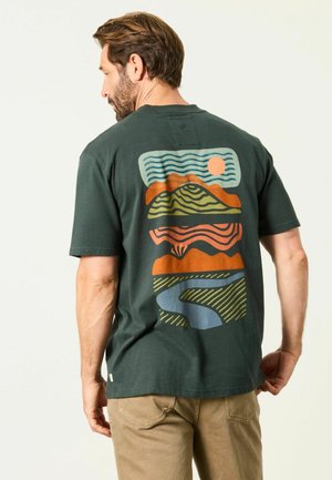 Man with brown hair and beard wears dark green t-shirt with colorful abstract landscape design on back, paired with beige pants.
