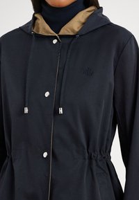 Tmavě modrá lehká bunda s béžovou kapucí, stříbrnými cvoky, nastavitelnými šňůrkami a elastickým pasem pro přizpůsobený vzhled.