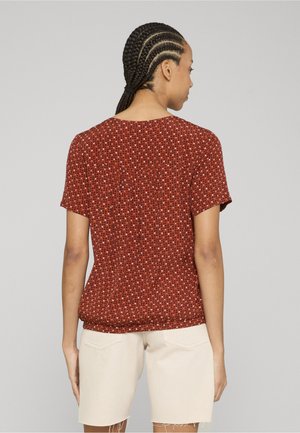 Žena s pletenicama, odjevena u crvenu majicu s kratkim rukavima s uzorkom i bež kratke hlače do koljena, stoji okrenuta leđima na jednobojnoj pozadini.