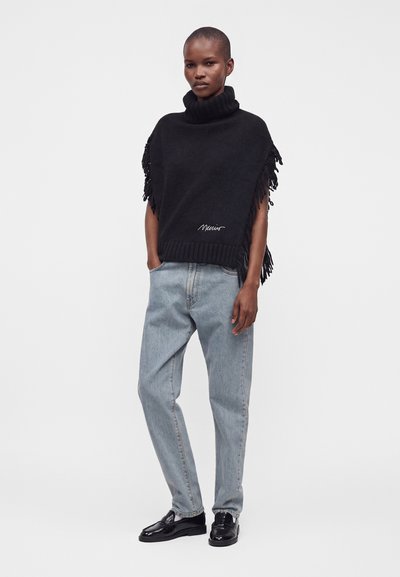 Femme portant un poncho en maille noire à franges avec col roulé, un jean bleu clair décontracté et des mocassins noirs brillants, debout les mains dans les poches.