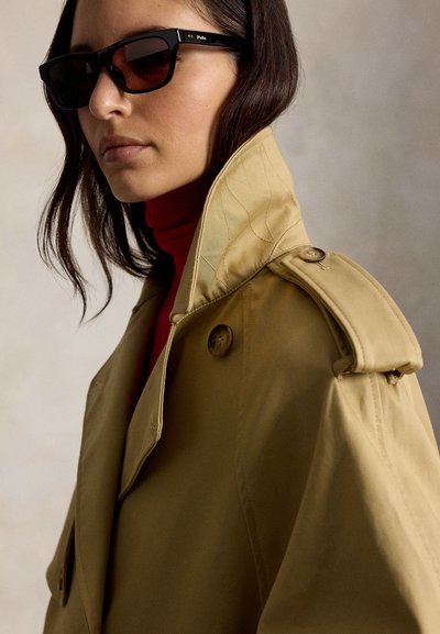 Γυναίκα που φοράει μαύρα γυαλιά ηλίου Polo, μπεζ παλτό τύπου τρεντς με ανασηκωμένο γιακά και κόκκινο πουλόβερ ζιβάγκο, μπροστά σε ουδέτερο φόντο.