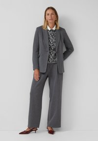 Vrouw staat tegen een muur in een grijs gestreept pak, een panterprint trui, een witte blouse, gouden oorbellen en bordeauxrode kitten heels.