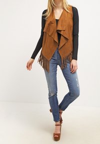 Chaleco de ante marrón con frente drapeado y detalles de flecos, usado sobre una camiseta negra y combinado con jeans azules desgastados y tacones plataforma marrones.