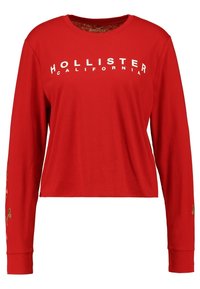 Röd långärmad tröja med texten "Hollister California" i vitt, prydd med dekorativa blommönster på ärmarna. Mjuk textur.