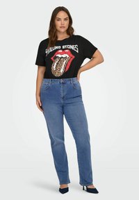 Czarna koszulka z czerwoną grafiką Rolling Stones i językiem w leopardzim wzorze, zestawiona z wysokim stanem niebieskimi dżinsami i spiczastymi szpilkami.