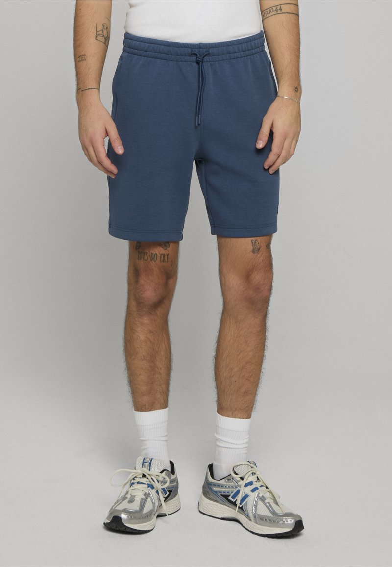 Blå bomullsshorts med elastisk midje og snor. Har sidelommer og en avslappet passform, kombinert med grå og blå joggesko.