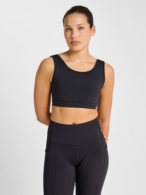 Ung kvinde med mørkt hår sat tilbage iført en sort sports-bh og sorte højtaljede leggings, stående mod en hvid baggrund.