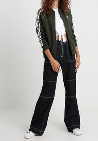 Grön bomberjacka med vita ränder, vit kortärmad skjorta, mörk denimbyxor med vida ben och kontrasterande sömmar, samt vita sneakers.