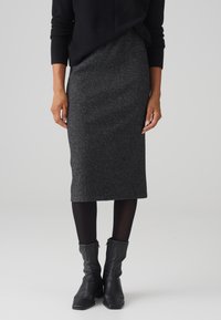 Femme portant une jupe midi côtelée gris foncé, des collants noirs, des bottines en cuir noir et un haut à manches longues noir sur un fond uni.