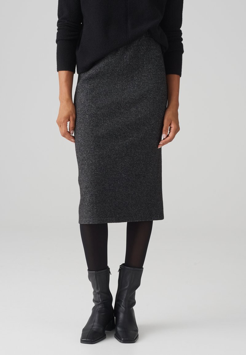 Femme portant une jupe midi côtelée gris foncé, des collants noirs, des bottines en cuir noir et un haut à manches longues noir sur un fond uni.