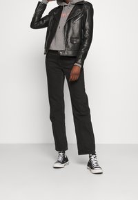 Giacca da motociclista in pelle nera, felpa grigia con cappuccio, jeans neri e sneakers alte nere. Presenta cerniere e dettagli con tasche.