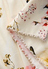 Mønstret stof med blomsterdesign i pink, gul og brun på en cremefarvet baggrund, med en flæset kant i lyserød.