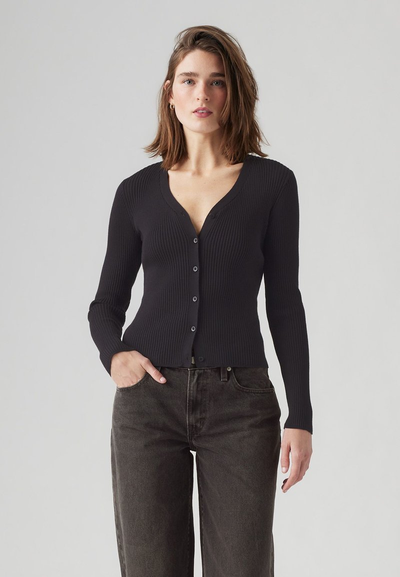 Schwarze gerippte Strickjacke mit V-Ausschnitt, Frontknöpfen und langen Ärmeln. Kombiniert mit dunklen, weit geschnittenen Hosen, die ein minimalistisches Design zeigen.