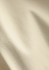 Krémová, textúrovaná tkanina s hladkým povrchom, predvádzajúca jemné záhyby a mäkké tiene, vyžarujúca ľahký vzhľad.