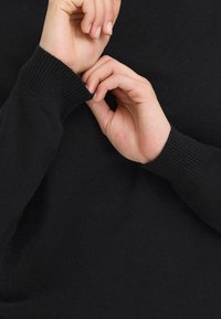 Zwarte gebreide trui met ribbelmanchetten. Handen zijn bezig de mouw aan te passen, waardoor de zachte textuur en de nauwsluitende pasvorm worden benadrukt.