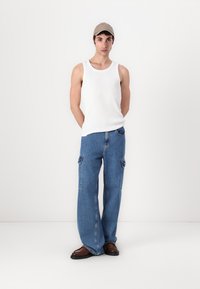 Witte mouwloze tanktop, blauwe wijde cargobroek met zijzakken en bruine schoenen; aangevuld met een beige pet. Eenvoudig, casual ontwerp.