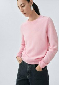 Suéter rosa claro de material suave con cuello redondo y puños acanalados, combinado con jeans oscuros. Presenta una silueta ajustada.