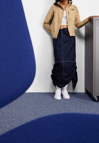 Beige zip-up jacka med sidofickor, ihopparad med en lång blå denimkjol. Vita sneakers med ett guldaccent. Grå arkivskåp i bakgrunden.