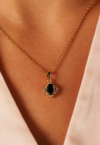 Collier en chaîne dorée avec un pendentif en pierre noire ovale entouré d'une bordure décorative de pierres précieuses transparentes.