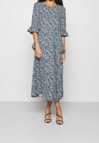 Blommig midiklänning i blått, vitt och grönt. Har volangärmar och en vågig design. Tillverkad av lätt tyg. Svart sandaler med remmar bärs.