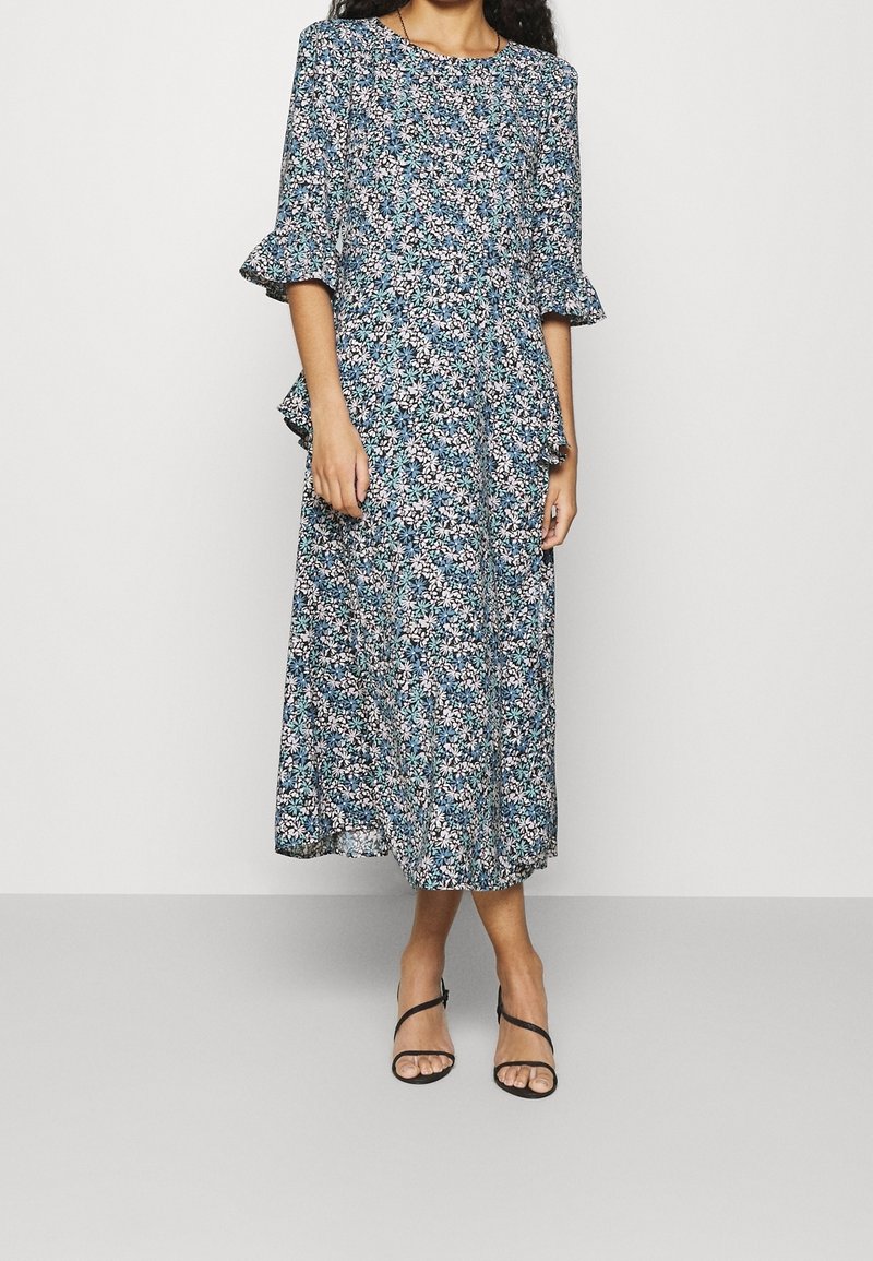 Blommig midiklänning i blått, vitt och grönt. Har volangärmar och en vågig design. Tillverkad av lätt tyg. Svart sandaler med remmar bärs.