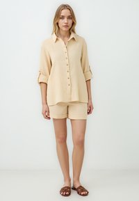 Camicia beige con bottoni e maniche arrotolate, abbinata a pantaloni corti coordinati. Tessuto strutturato, vestibilità comoda e design minimalista. Sandali a scivolo marroni.