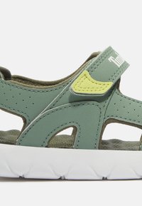 Sandalia infantil verde y amarilla con tiras ajustables y suela blanca texturizada mostrada en vista lateral de cerca.